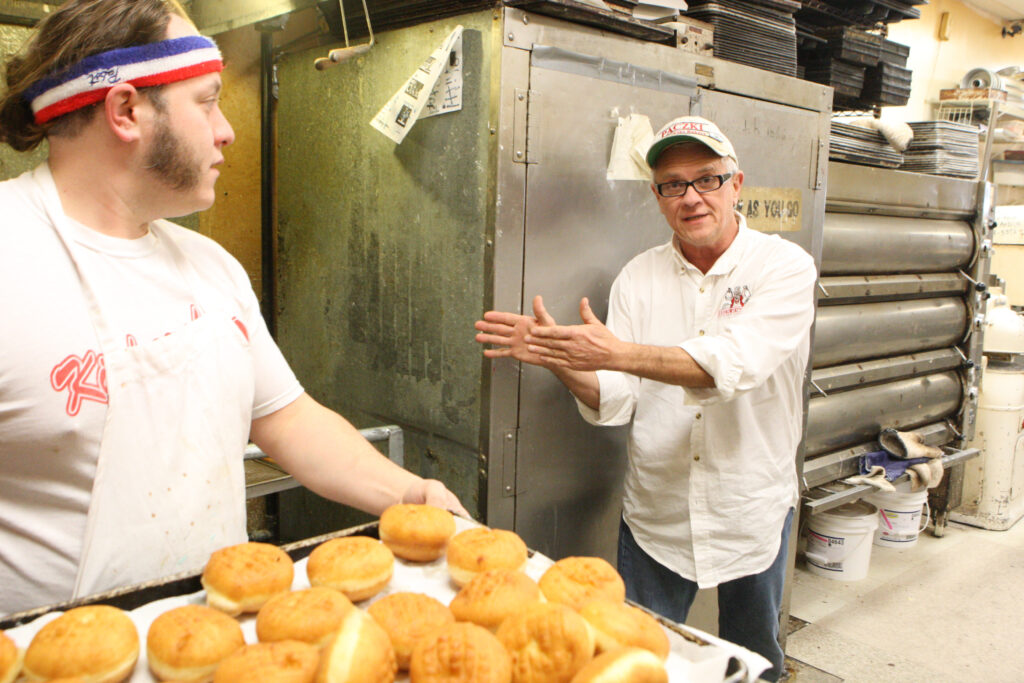 Tim Kiedrowski, owner of Kiedrowski's Simply Delicious Bakery, explains the process of making Polish-style doughnuts, known as "paczki," Jan. 18 as his son Michael moves a rack of the treats for cooling. The doughnuts are a tradition in the days before Lent. (CNS photo/Dennis Sadowski)
